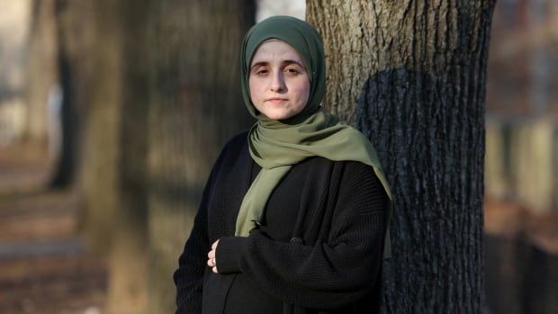 A pregnant woman wearing a green hijab and dressed in black stands in front of a tree holding her belly