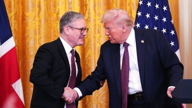 Two men, flanked by British and U.S. flags, shake hands while smiling.