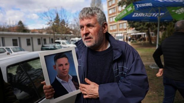 A bearded, grey-haired man in a coat outdoors holds up a framed photo of a younger man.