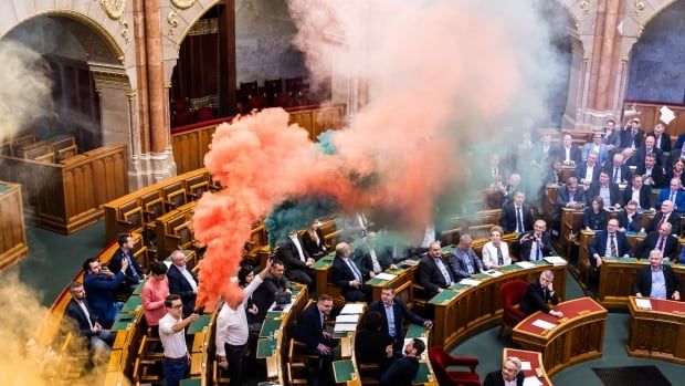 Large clouds of smoke, orange and green in colour, hang in the air inside what appears to get a government chamber, with dozens of people sitting or standing.