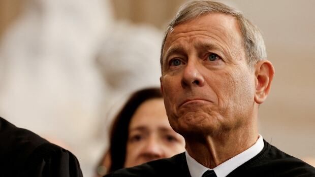 A white-haired cleanshaven man has a pout on his face in a closeup photo. He is wearing a tie, collared shirt and a judicial robe.