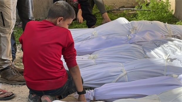 A boy sits next to bodies wrapped in white shroud.