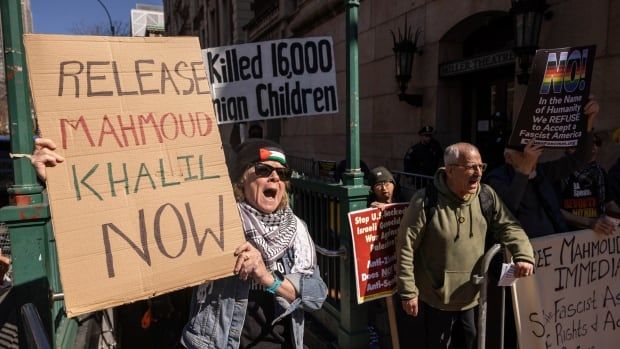 Protesters hold signs and yell slogans. A protester in the front is holding a large cardboard sign that says "Release Mahmoud Khalil now". 