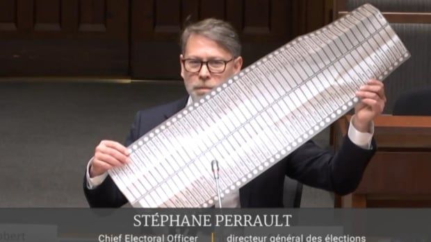 A man sits in front of a microphone and holds up a ballot that is nearly a metre in lenght.