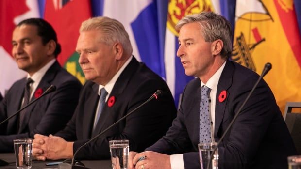 Three men wearing suits sit at a long table with microphones in front of them. Provincial flags can be seen behind them.