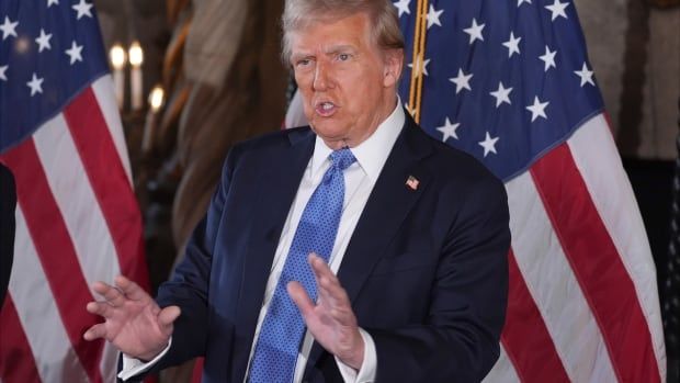 An older clean shaven man in a suit and tie is shown speaking in front of American flags.