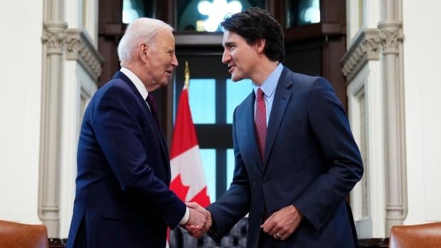 U.S. President Joe Biden shakes hands with Prime Minister Justin Trudeau inside Parliament in Ottawa. A Canadian flag stands between them in the background. 