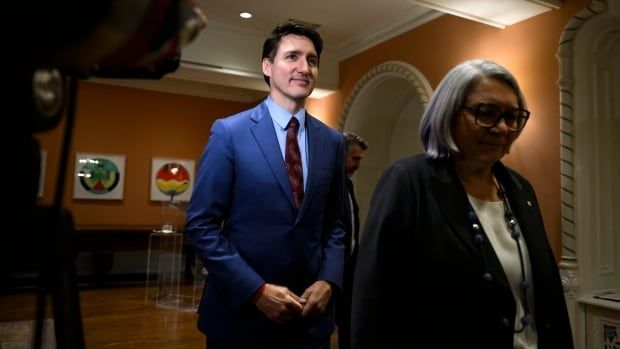 A brown-haired man in a blue suit and a grey-haired woman in a dark suit and glasses walk through the hallway of a government building.