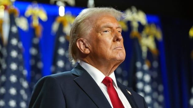 A man in a dark suit and red tie is seen on stage in front of several U.S. flags.