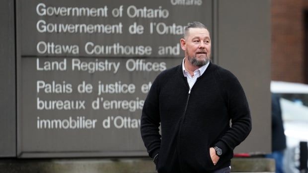 A man in a zip-up sweater and collared shirt walks near a courthouse in autumn.