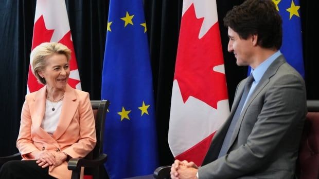 Ursula von der Leyen sits next to Prime Minister Justin Trudeau, both on chairs in front of European Union and Canadian flags.