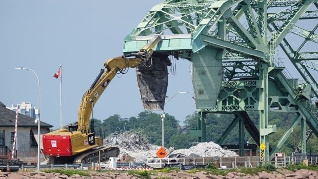 A large piece of construction equipment drives halfway up a pile of rubble in front of  heavily-damaged green metal bridge. 