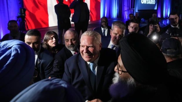 A man in a suit and blue tie greets a crowd of supporters. A Canadian flag is in the background.