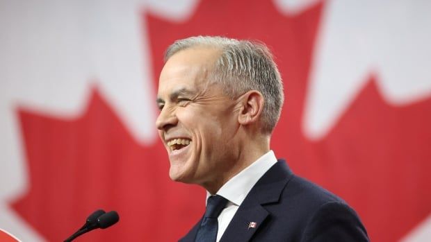 A person smiles while speaking in front of a large Canadian flag.