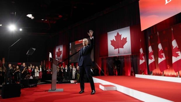 A man with grey hair and a dark suit waves to a crowd of supporters. Canadian flags are visible.