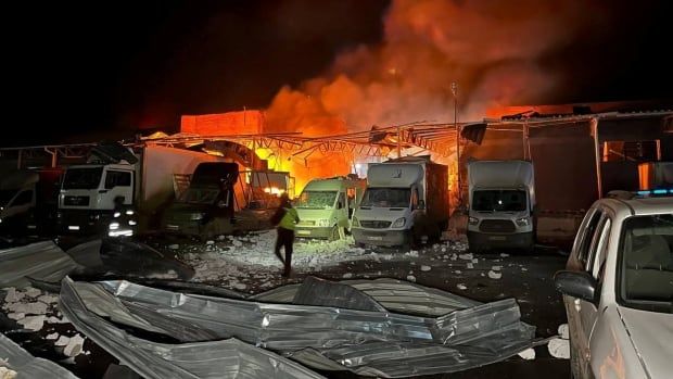 Vehicles are shown lit up by a background of fire in a parking lot.