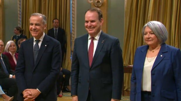 A man in a navy blue business suit stands between another man in a dark suit and a woman in a blue business suit. 