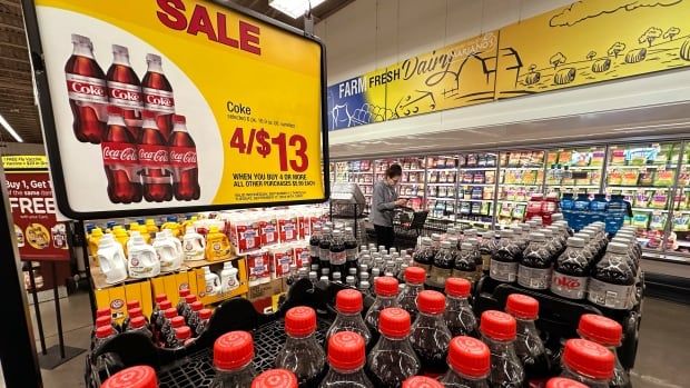 A pallet of plastic Coke brand bottles with red caps is visible underneath a "SALE" sign in a grocery store. A person is visible shopping in front of a wall of freezers in the background. 