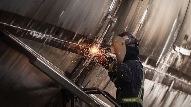 Sparks fly as a worker applies a tool to the curved side of a very large object, which appears to be made of metal. The worker is wearing a large welding mask and helmet and is dressed in dark clothes. 