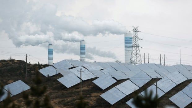 Smoke rises from three chimneys in the background. In the foreground, there are rows of solar panels.