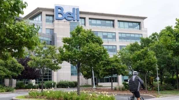 The outside of a building is shown, with big blue letters saying Bell at the top. A man is biking past at the bottom of the image. 