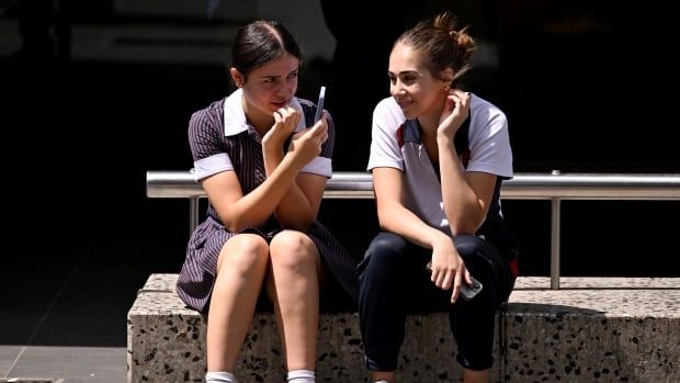 Two girls who appear to be teens are shown sitting on a step, with one holding up a phone.