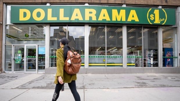 The front of a store is visible, with the word DOLLARAMA emblazoned in big yellow letters on a green banner above the store's windows. A person is walking down the street in front of it, their face turned away from the camera, wearing a brown backpack and a baseball hat. 