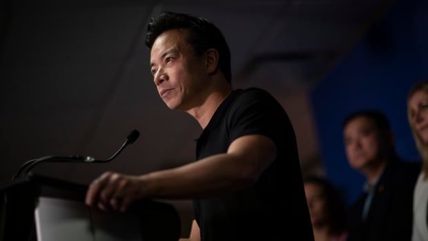 An man with short black hair wearing a black fitted T-shirt speaks at a podium, with a few other people behind him.