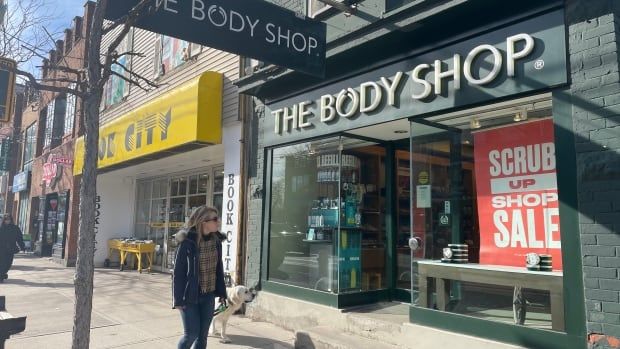 A woman and her dog walk by a storefront with a sign reading The Body Shop.