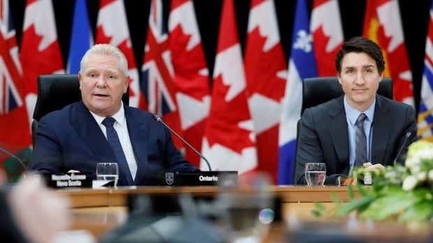 Two men sit at a table, behind which are several Canadian and provincial flags. 