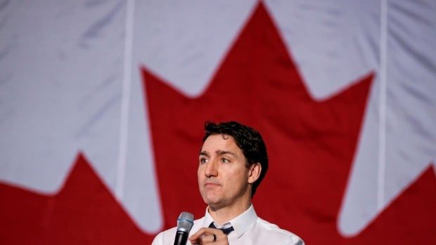 Justin Trudeau standing in front of a Canadian flag, holding a mic.