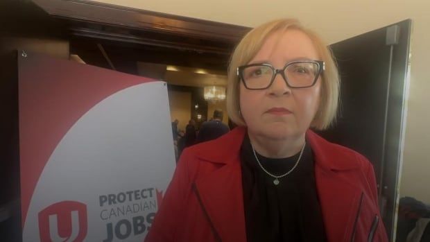 A woman looks at the camera. Behind her is a blurry ballroom and a sign that says "protect Canadian workers." 