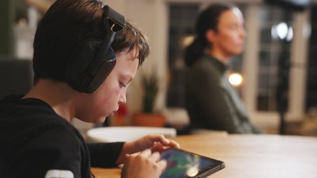 A boy, wearing headphones, sitting at a table, playing on a tablet. 