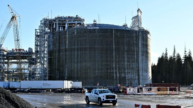 A white pickup truck drives on a road next to a liquid natural gas storage tank. 