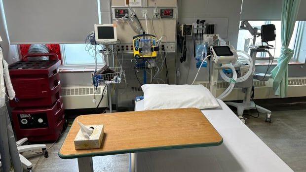 An empty hospital bed with white sheets and a variety of medical equipment arranged at the head of it.