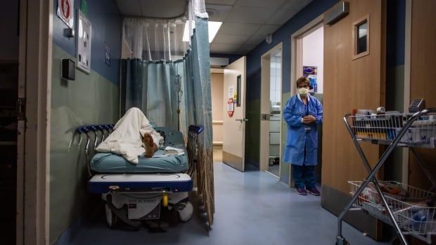 A patient rests in a corridor waiting for a room in an emergency room. A curtain is partly drawn around them as a health-care worker looks on.