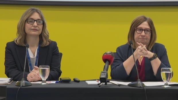 Two women with black blazers sit behind table with microphones