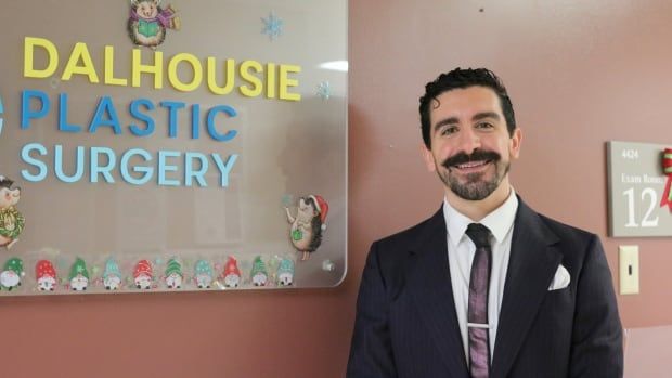 A smiling man with slicked back hair, a mustache and a beard wears a suit and a tie. He stands next to a sign saying "Dalhousie Plastic Surgery."