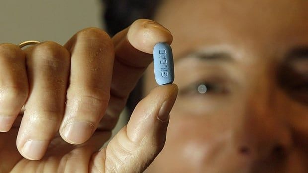 Someone holds up a blue HIV treatment pill between their fingers. In the background you can partially see the face of the person holding the pill. 