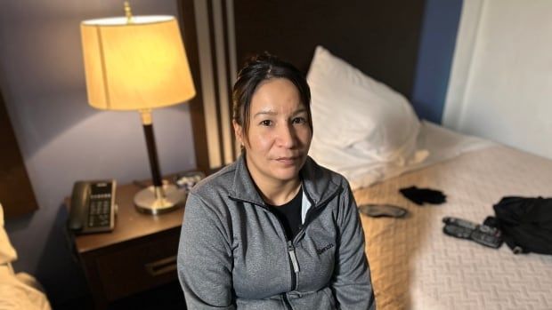 A woman in a grey zip-up hoody sits on a bed in a hotel room.