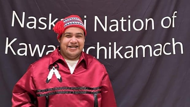 A man smiles at the camera, wearing a read collared shirt. He stands in front of a flag that says "Naskapi Nation of Kawawachikamach."