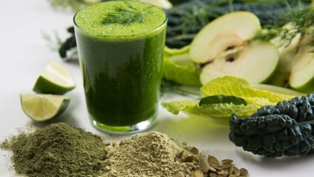 Lime, green powder, sunflower seeds, kale, lettuce and green apples are arranged on a table. At the centre of the arrangement, there's a glass of green juice. 