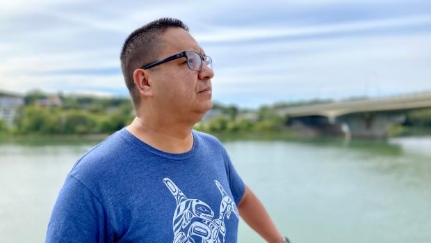 A man in dark glasses and with black hair stares into the distance with the river seen behind him. 