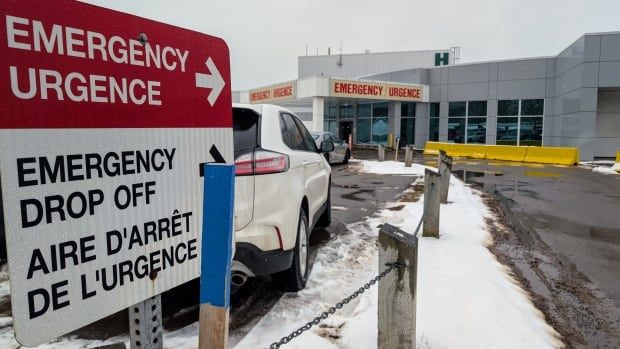 Emergency directional sign in parking lot, in foreground, with hospital EMERGENCY department and sign in background, in winter.