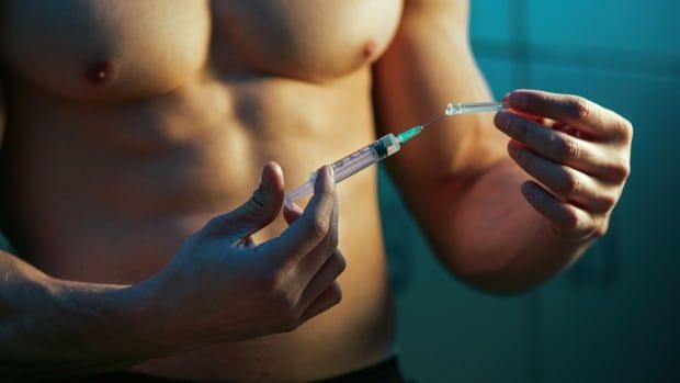 A shirtless muscular man removes the cap from a needle.