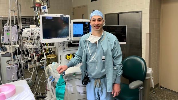 A man in medical scrubs stands next to anesthesia equipment.