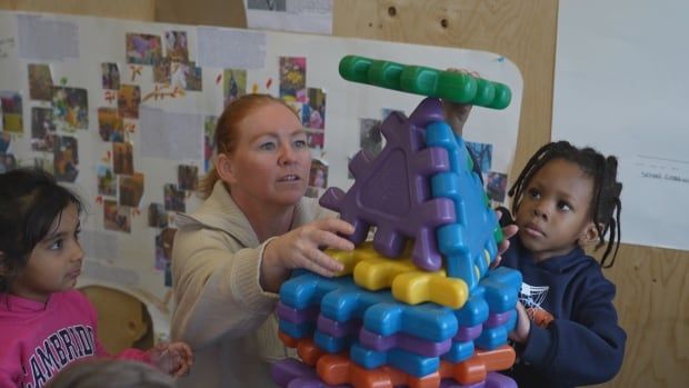 A female educator holds colourful, oversized waffle block tiles steady as a preschool-aged boy builds a tower with them. A preschool-aged girl looks on from the left.