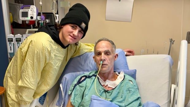 a teenager in a yellow hospital robe stands next to a man in a bed with a bandaged arm