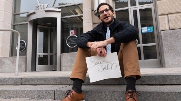 A man sits on the steps outside a building holding a small sign that says "Save USAid."