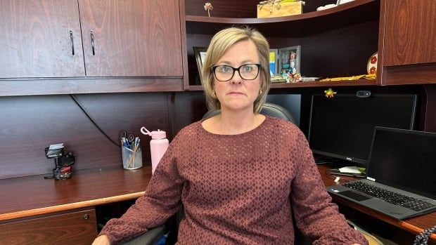 A woman with a blond bob wearing a terracotta blouse sits in an office chair just behind a large desk. She has a serious expression